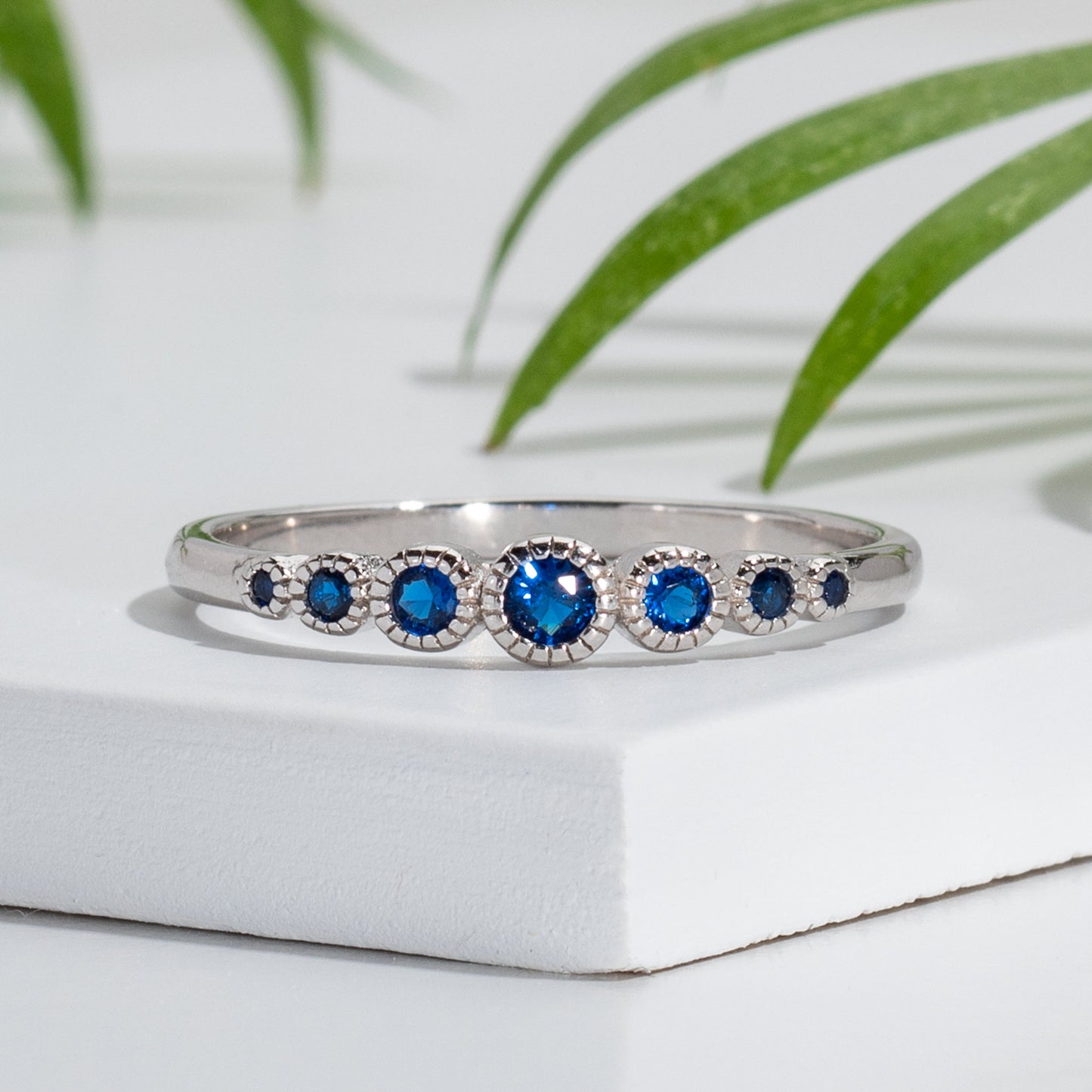 A sterling silver bezel round-cut ring with a vibrant blue cubic zirconia stone set on a white surface, surrounded by green foliage.