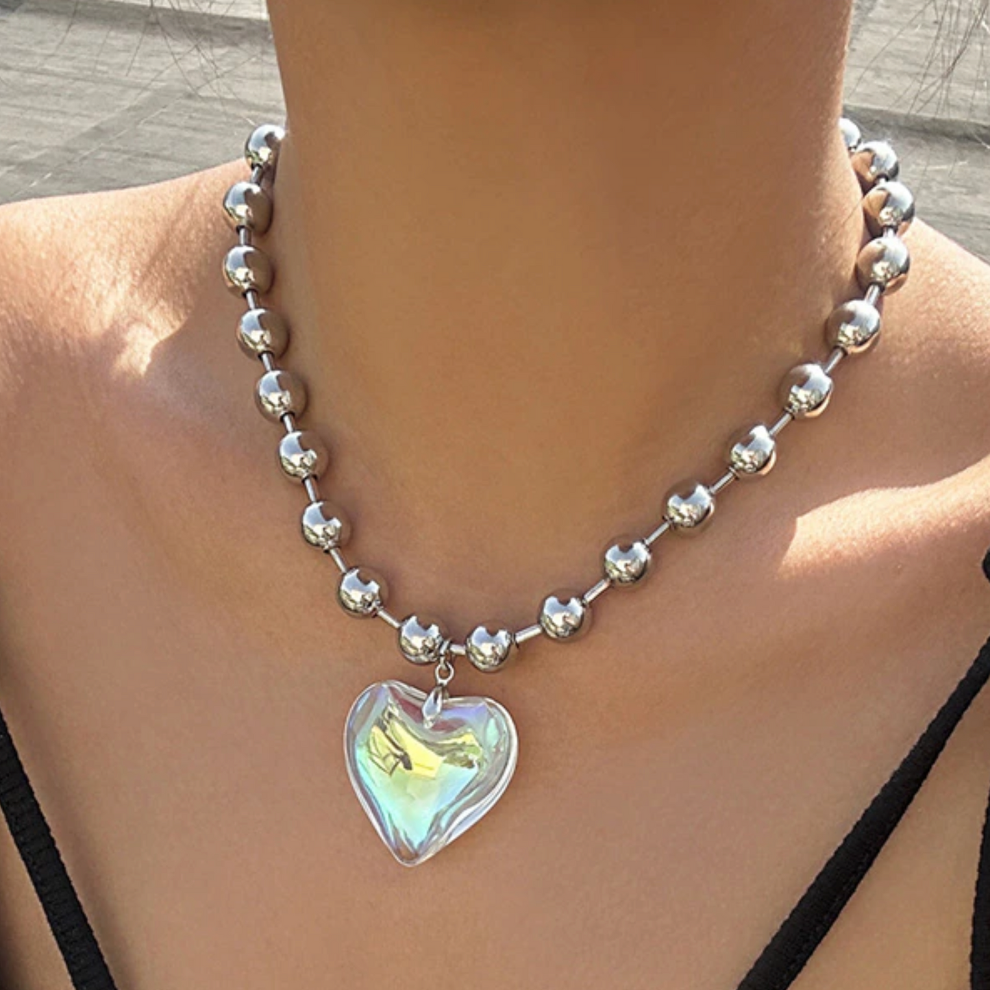 A close-up image of a person wearing a silvertone ball chain necklace with a green iridescent crystal heart pendant.