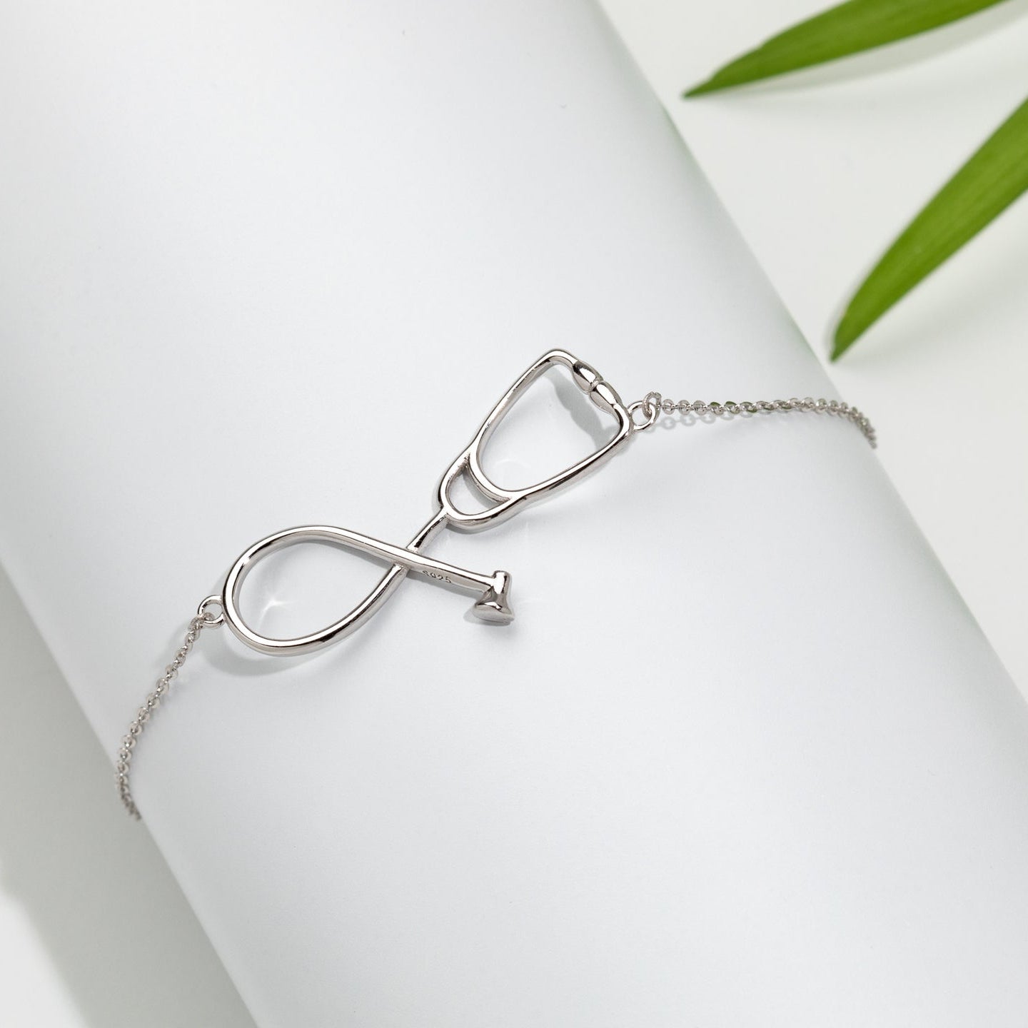 A sterling silver stethoscope bracelet with a double-loop chain design, displayed on a white background next to green foliage.