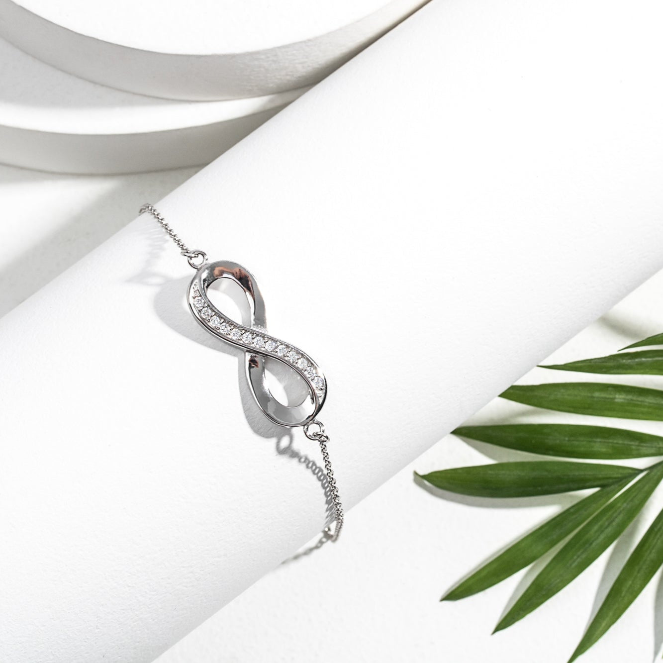 A sterling silver cubic zirconia infinity bracelet displayed on a rolled-up white paper, with a green leaf and a white background.