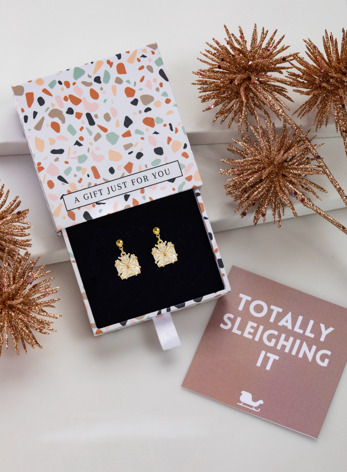 Gold-tone crystal floral earrings in a patterned gift box beside a pink card