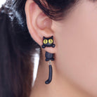 Black cat earring with yellow eyes and dangling tail, worn on an ear