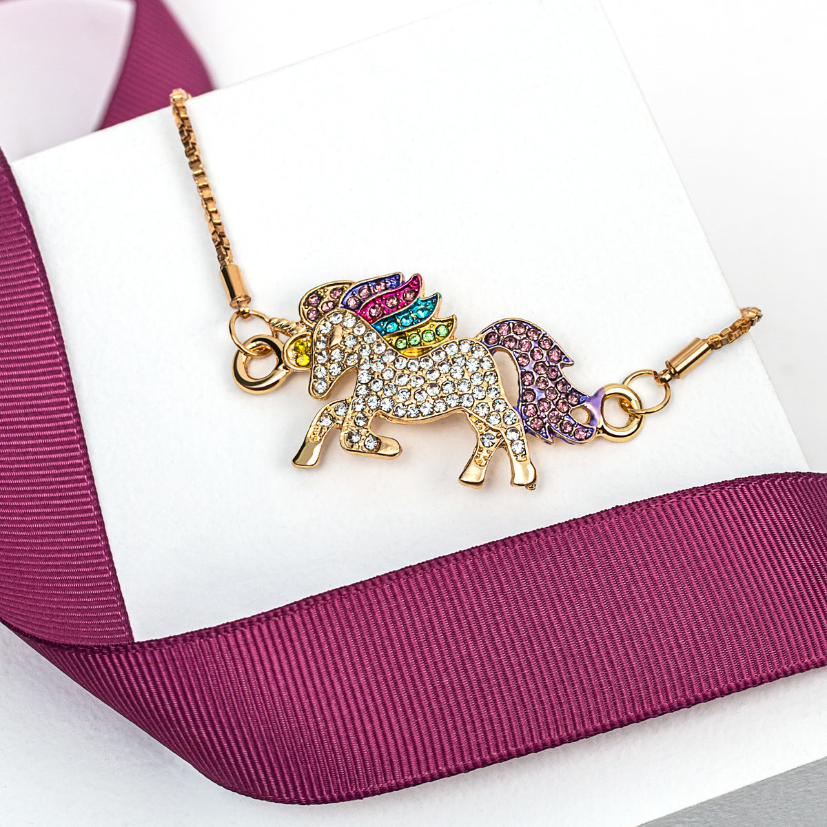 Goldtone crystal unicorn bracelet displayed on a white cushion beside a pink ribbon